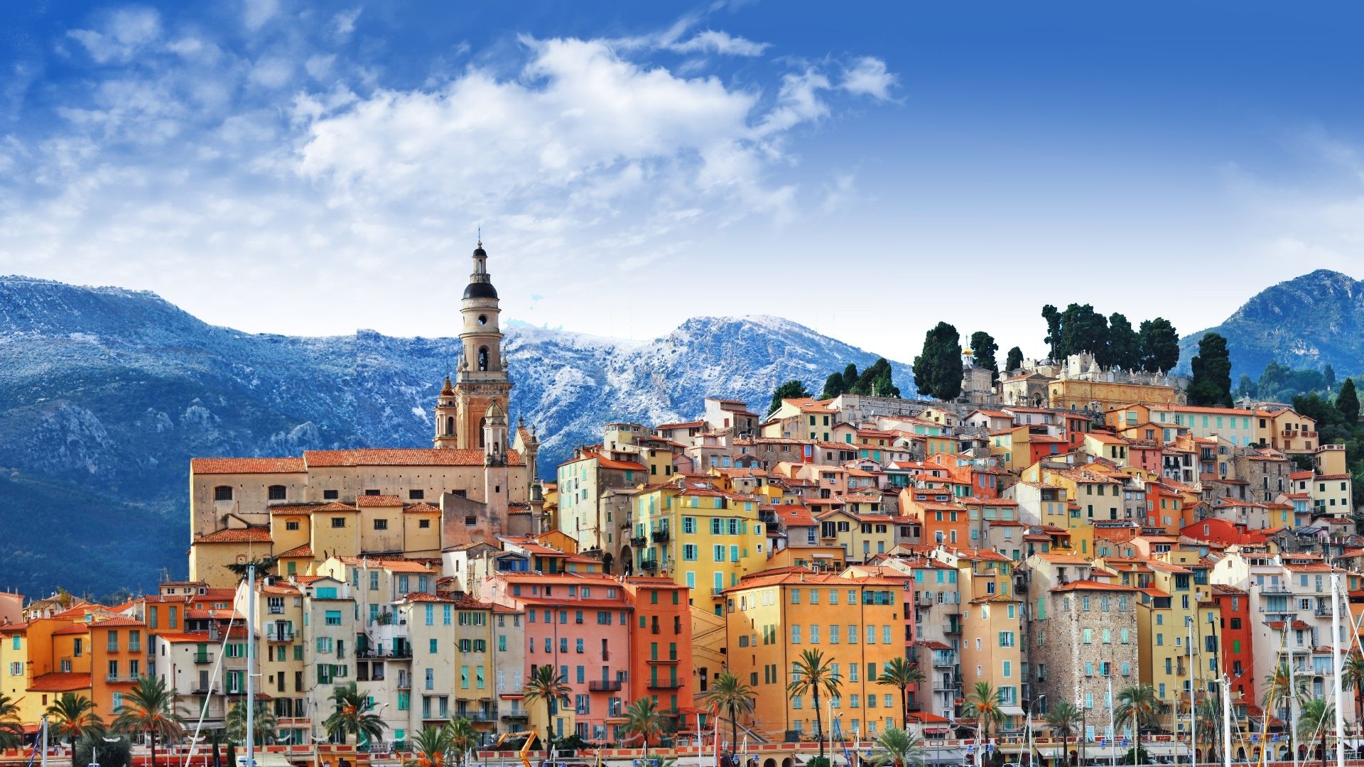 Menton's pastel old town from the sea — the Basilica of Saint-Michel-Archange on the headland marks the eastern French border. The Cocteau Museum (the Bastion on the harbor, painted by Cocteau himself in 1957) is on the quay; the Jean Cocteau Museum proper, the larger 2011 building, sits two hundred meters east. The Lemon Festival every February closes the streets for two weeks.