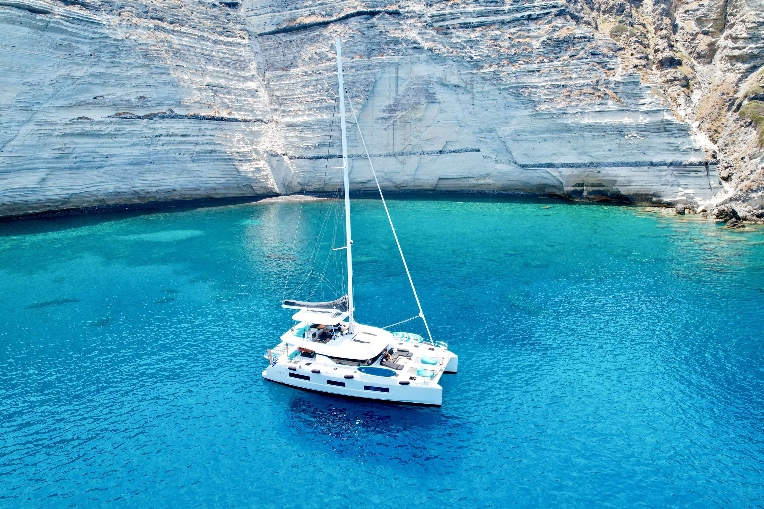 Crewed catamaran at anchor in a limestone-cliff Mediterranean cove, aerial view